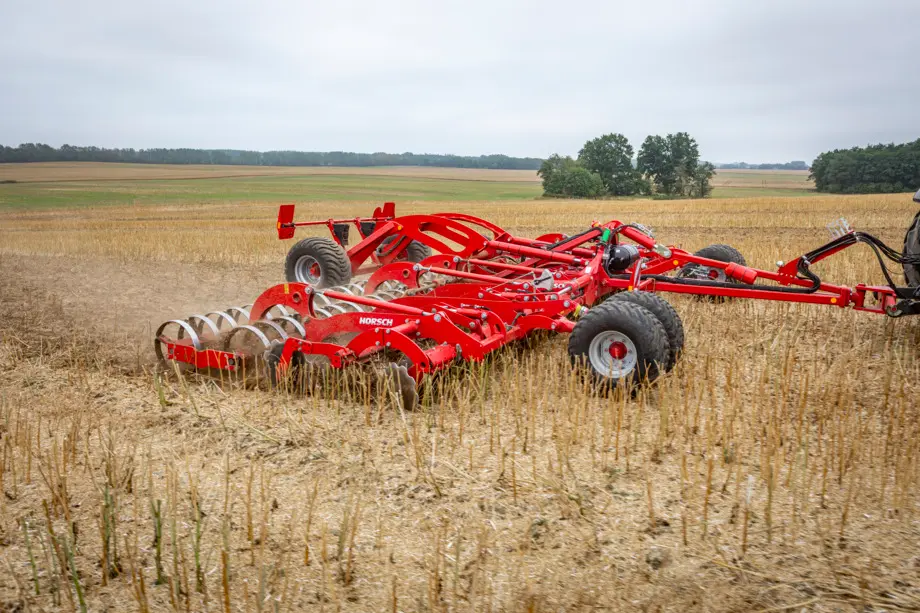Brona talerzowa Horsch Joker RT w trakcie uprawy pola po zbiorach. Widok z boku na rzędy talerzy tnących oraz tylny wał pierścieniowy zagęszczający glebę. W tle rozpościera się pagórkowaty krajobraz rolniczy pod zachmurzonym niebem.