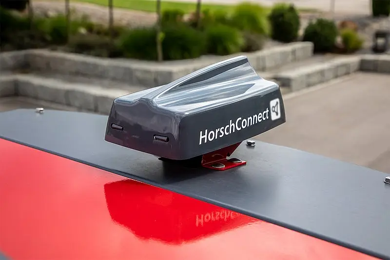 Zbliżenie na szary, opływowy moduł anteny telemetrycznej z białym napisem "HorschConnect", przykręcony do czerwonej, metalowej obudowy maszyny rolniczej.