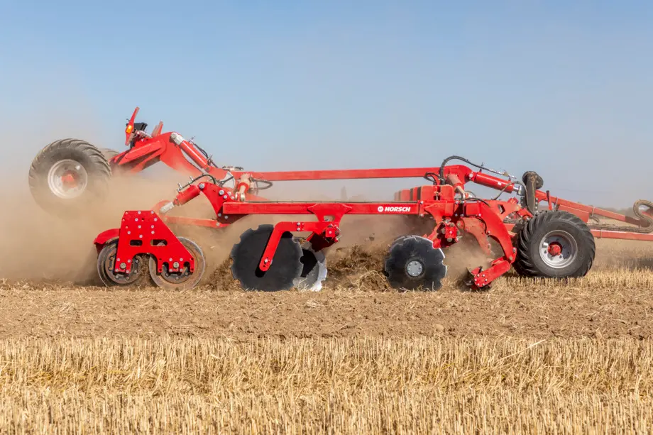 Czerwona brona talerzowa Horsch Joker HD podczas intensywnej pracy na suchym ściernisku. Widoczne są duże, ząbowane talerze wgryzające się w ziemię oraz uniesione koła transportowe. Maszyna wzbija tumany kurzu na tle czystego nieba.
