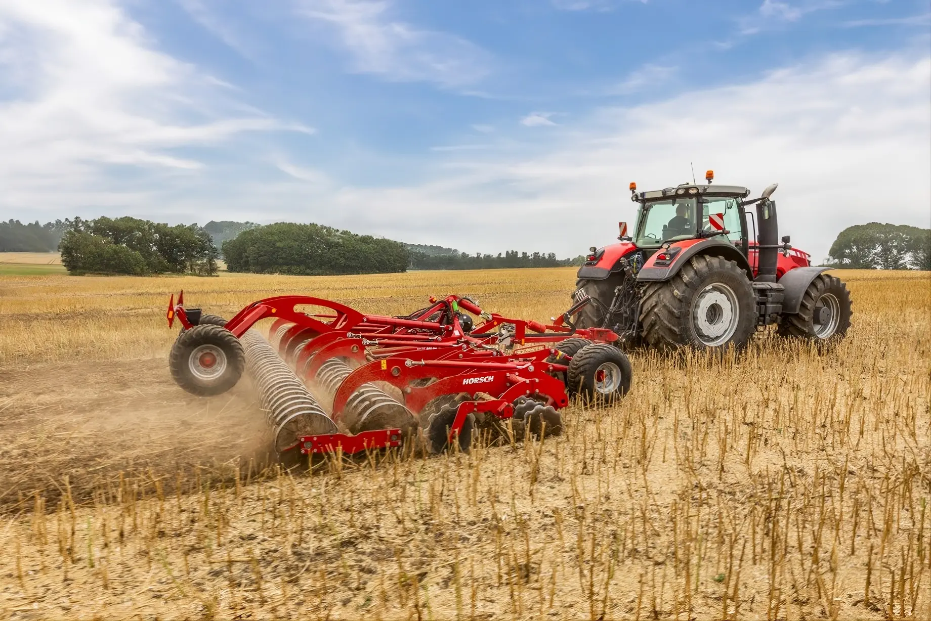 Duży czerwony ciągnik rolniczy ciągnący bronę talerzową Horsch Joker RT przez ściernisko. Dynamiczne ujęcie maszyny pracującej w pełnym słońcu, wzbijającej pył podczas doprawiania gleby. W oddali widać pasy zieleni i błękitne niebo z lekkimi chmurami.