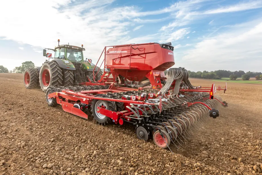 Czerwony, szeroki siewnik Horsch Pronto ciągnięty przez zielony ciągnik rolniczy na zaoranym polu pod błękitnym niebem z delikatnymi chmurami.
