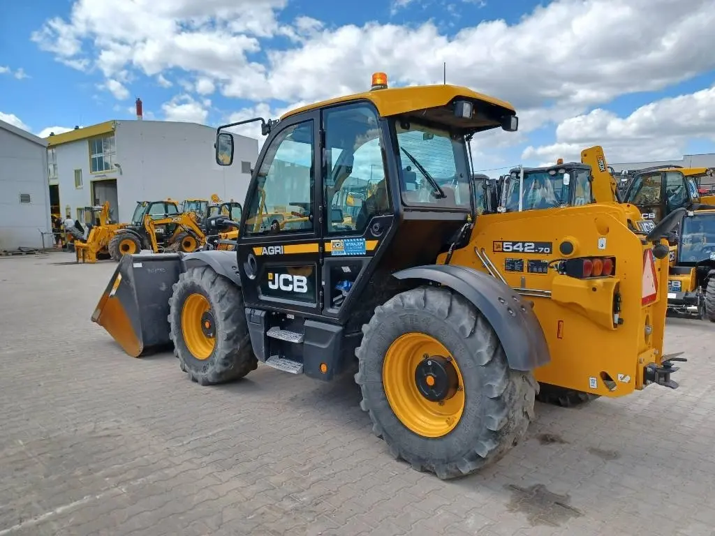 Profil ładowarki teleskopowej JCB 542-70 Agri stojącej na brukowanym placu handlowym. Widoczna kabina operatora, detale tylnego mostu oraz inne maszyny budowlane w tle pod błękitnym niebem