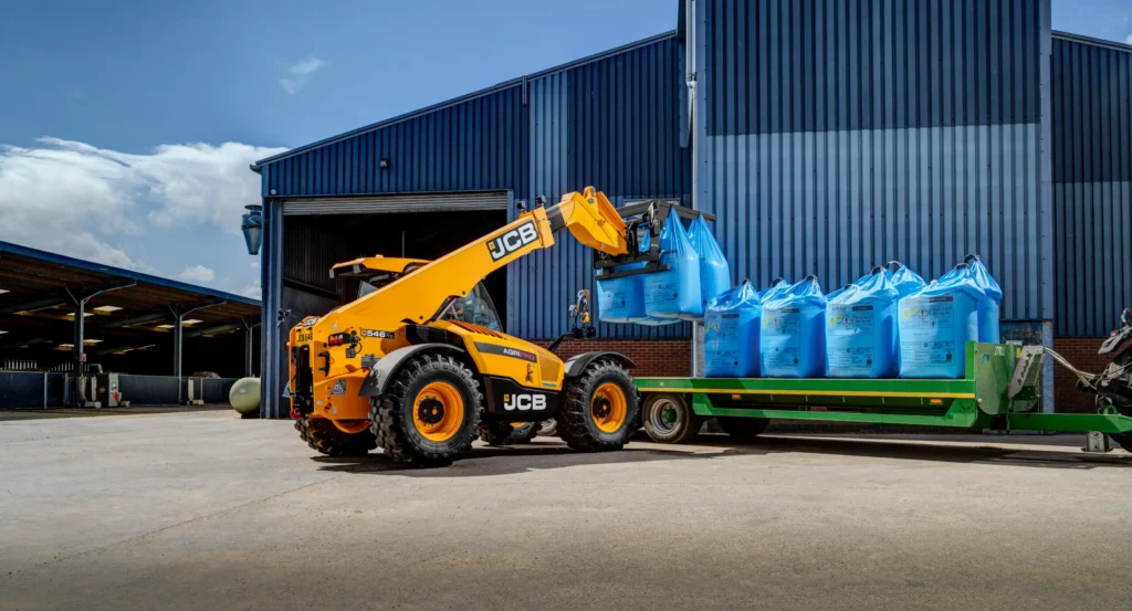 Ładowarka teleskopowa JCB 546-70 Agri Pro ładuje niebieskie worki typu Big-Bag na zieloną przyczepę transportową przed nowoczesną niebieską halą magazynową.