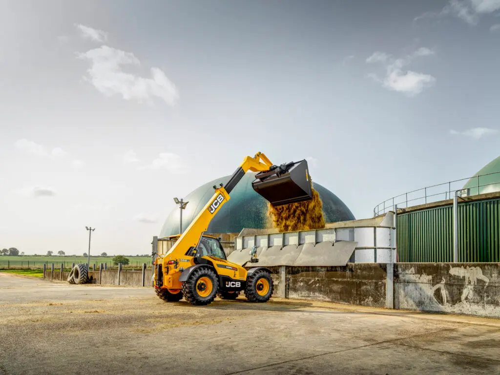 Żółta ładowarka teleskopowa JCB 555-70 Agri Pro z uniesionym wysięgnikiem, wysypująca ziarno z łyżki do betonowego silosu na terenie gospodarstwa. W tle widoczna zielona kopuła biogazowni.